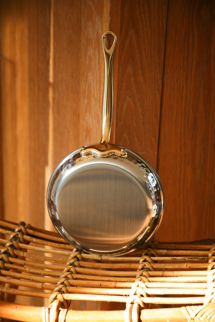 Mauviel M'ELITE B hammered 5-ply frying pan set with brass handles, shown in a lifestyle kitchen setting.